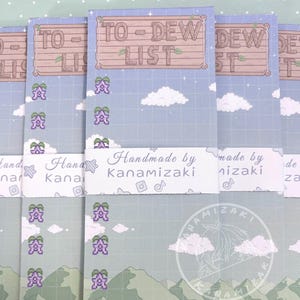 May include: A stack of "To-Dew List" notepads. Each notepad features a light blue sky background with white clouds, pixelated purple flowers, and green mountains. The top of each notepad has the words "To-Dew List" in a wooden sign design.