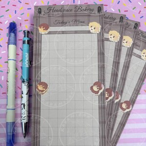 May include: A stack of gray "Headspace Bakery Today's Menu" notepads with grid lines, decorated with cartoon character stickers. A blue pen and a blue and white "Moomin" pencil are on the left.
