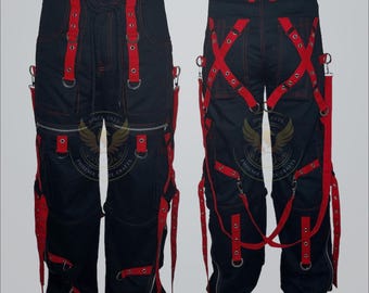 Op de afbeelding: Zwarte broek met rode bandjes en accenten. De broek heeft meerdere zakken, metalen ringen en een ritssluiting. Het ontwerp omvat rode bandjes die kruisen en langs de zijkanten lopen, wat een unieke, edgy look creëert. De broek heeft een knoopsluiting.