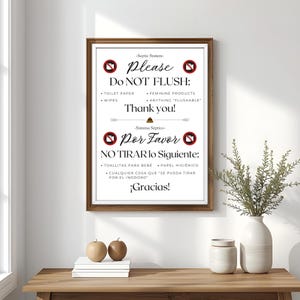 May include: A framed sign with text in English and Spanish, advising against flushing certain items. The sign has a brown frame and is displayed on a wooden table. Decorative elements include a vase with greenery and two decorative apples.