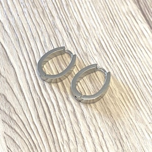Könnte beinhalten: Ein Paar silberfarbene ovale Creolen. Die Ohrringe sind aus glänzendem Metall gefertigt und haben ein schlichtes, modernes Design. Sie werden auf einer hellen Holzoberfläche präsentiert.