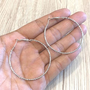 May include: A pair of silver hoop earrings with a textured pattern. The earrings are circular and have a simple, elegant design. The hoops are held in a hand, showing their size and style.