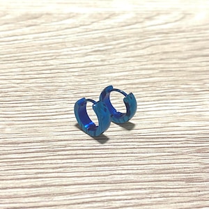 May include: Two small, blue hoop earrings with a textured surface. The earrings are made of metal and have a simple, modern design. They are displayed on a light-colored wooden surface.