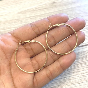May include: A pair of gold-coloured hoop earrings. The earrings are round and have a simple, classic design. The hoops are held in a hand, and the background is a light wood grain.