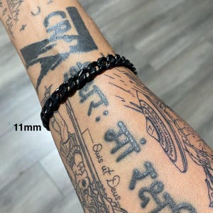 Puede incluir: Una pulsera de cadena negra, de 11 mm de ancho, se muestra en un brazo con tatuajes. Los eslabones de la pulsera están claramente definidos, y la estética general es moderna y audaz. La pulsera es un accesorio elegante.