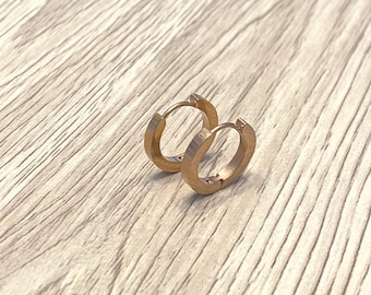 Könnte beinhalten: Ein Paar kleine, roségoldene Creolen. Die Ohrringe bestehen aus einem glatten, metallischen Material und haben ein schlichtes, minimalistisches Design. Sie liegen auf einer hellen Holzoberfläche.