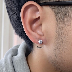 Può includere: Primo piano di un orecchio con un singolo orecchino a bottone rotondo e trasparente. L'orecchino ha un diametro di 6 mm ed è incastonato in metallo color argento. La persona indossa una felpa con cappuccio grigia.