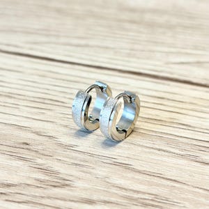 Peut inclure: Une paire de boucles d'oreilles créoles argentées avec une bande texturée et scintillante. Les boucles d'oreilles sont en métal brillant et sont présentées sur une surface en bois brun clair.