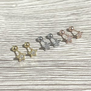 May include: A collection of star-shaped stud earrings in gold, silver, and rose gold tones. Each earring features a small, round stud and a dangling star embellished with small, clear gemstones. The earrings are displayed on a light wood-grain surface.