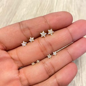 May include: Several gold-coloured star-shaped stud earrings. The earrings are of varying sizes and feature clear, sparkling accents. The studs are displayed on a light-coloured surface, showcasing their delicate design and shine.