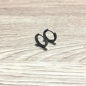 Peut inclure: Deux petites boucles d'oreilles créoles noires. Les boucles d'oreilles sont en métal foncé et ont un design simple et minimaliste. Elles reposent sur une surface en bois brun clair.