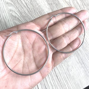 Puede incluir: Dos pendientes de aro plateados. Los pendientes son redondos y tienen un diseño simple y minimalista. Los aros se sostienen en la mano de una persona, y el fondo es una superficie de madera de color claro.