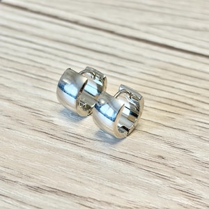 Puede incluir: Dos pendientes de aro plateados con una superficie brillante y reflectante. Los pendientes son gruesos y tienen un diseño sencillo y moderno. Están sobre una superficie de madera clara.