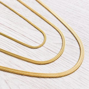 Peut inclure: Trois colliers en chaîne dorés en chevrons de différentes épaisseurs sont présentés sur un fond clair en grain de bois. Les chaînes sont disposées en forme incurvée, mettant en valeur leur texture et leur design. Les colliers sont un bijou classique.