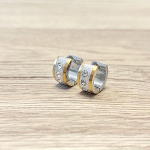 Peut inclure: Une paire de créoles argentées avec des détails dorés. Chaque boucle d'oreille présente une bande de strass scintillants et une bande dorée. Les boucles d'oreilles sont posées sur une surface en bois brun clair.
