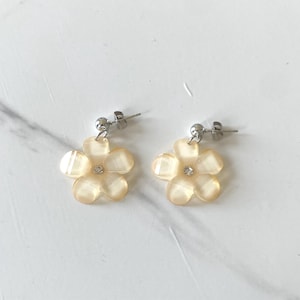Puede incluir: Un par de pendientes con forma de flor. Cada pendiente presenta una flor translúcida de color amarillo claro con cinco pétalos y una pequeña gema transparente en el centro. Los pendientes tienen postes y respaldos plateados.