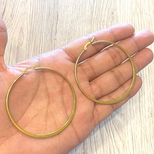 May include: Two gold-coloured hoop earrings. The earrings are circular with a flat, polished surface. The earrings are shown against a light-coloured background.