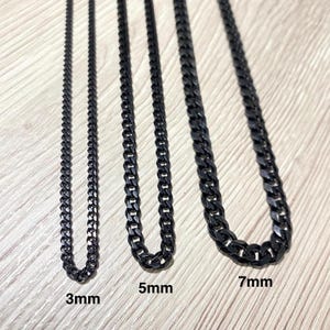 May include: Close-up of three black chain necklaces in varying widths: 3mm, 5mm, and 7mm. The necklaces are displayed on a light-colored wooden surface. The chains have a curb link design.