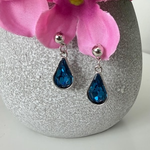 Peut inclure: Paire de boucles d'oreilles pendantes argentées avec des pierres en forme de goutte d'eau, de couleur bleu sarcelle. Les boucles d'oreilles sont présentées sur un fond gris texturé, avec des fleurs roses.