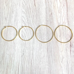 May include: Four gold-coloured hoop earrings of varying sizes are displayed on a light wood-grain surface. The earrings are arranged in a row, showcasing their circular design and shiny finish. The earrings are made of a thin metal.