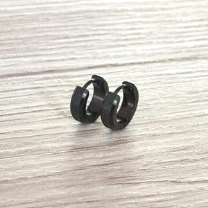 Peut inclure: Une paire de boucles d'oreilles créoles noires avec une finition texturée. Les boucles d'oreilles sont petites et rondes, avec un design simple et moderne. Elles sont présentées sur une surface claire imitation bois.