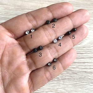 Pode incluir: Uma coleção de seis pares de brincos de pressão em vários estilos. Os brincos apresentam pedras preciosas pretas e transparentes, alguns com um design redondo e facetado. Os brincos são exibidos em uma superfície clara.