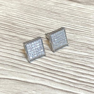 Peut inclure: Une paire de boucles d'oreilles carrées. Chaque boucle d'oreille est de couleur argentée et incrustée de petites pierres scintillantes. Les boucles d'oreilles sont présentées sur une surface en bois clair.