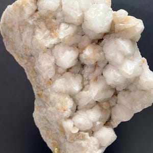 May include: A cluster of white quartz crystals with a rough, textured surface. The crystals are clustered together, creating a bumpy, irregular shape. The base of the quartz has a light tan color. The quartz is displayed on a stand with gold-colored supports.