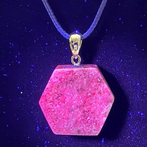 May include: A hexagonal pendant with a vibrant pink hue, suspended from a black cord. The pendant is attached to a gold-colored bail. The background is a dark blue color.