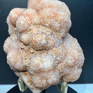 May include: A close-up of a natural mineral specimen. The rock has a rough, textured surface with a mix of peach, tan, and white colours. The mineral is mounted on a clear stand against a dark background.