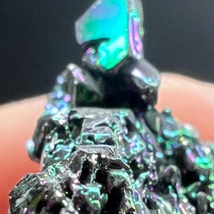 May include: Close-up of a mineral specimen with a dark, textured surface. The mineral exhibits iridescent colors, including shades of green, blue, and purple, creating a shimmering effect. The specimen has a crystalline structure.