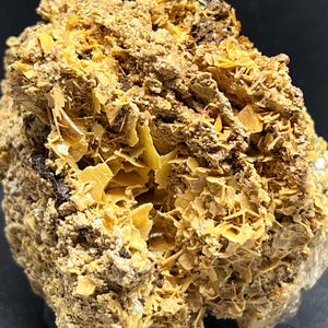 May include: A close-up of a mineral specimen with a rough, textured surface. The rock is primarily tan and brown, with patches of bright yellow, crystalline formations. The overall appearance is rugged and natural.