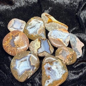 May include: A collection of polished agate stones in various shapes and sizes. The stones display a range of colors, including brown, white, blue, and orange, with intricate patterns and crystal formations. The agates are arranged on a dark, textured surface.