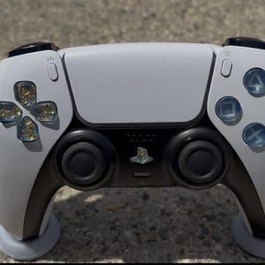 May include: A white and black PlayStation 5 controller. The controller has a white faceplate with black accents, including the analog sticks and triggers. The buttons have a blue hue. The D-pad has a gold glitter design.