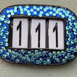 May include: A black, rectangular house number plaque with the number "111" displayed on white backgrounds. The plaque is framed with a border of blue and turquoise rhinestones.