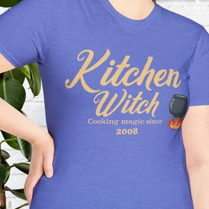 May include: A blue t-shirt with the words "Kitchen Witch" in gold script, along with "Cooking magic since 2008." A cauldron graphic is on the right. The shirt is a solid color and has a crew neck.