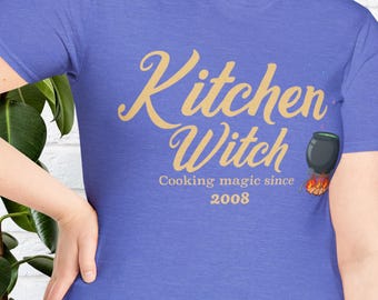 May include: A blue t-shirt with the words "Kitchen Witch" in gold script, along with "Cooking magic since 2008." A cauldron graphic is on the right. The shirt is a solid color and has a crew neck.