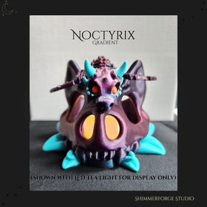 May include: A purple and teal dragon skull-shaped candle holder. The candle holder has two eye-shaped openings and a dragon figure on top. The text "NOCTYRIX GRADIENT" is at the top. The text "SHIMMERFORGE STUDIO" is at the bottom.