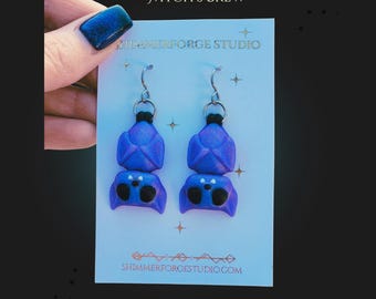 May include: A pair of handmade blue earrings with a unique design, featuring a top section and a lower section resembling a game controller. The earrings are displayed on a white card with the text "Witch's Brew" and "SHIMMERFORGE STUDIO".