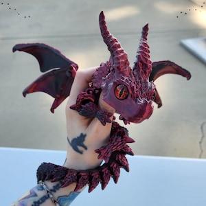 May include: A detailed, burgundy-colored dragon figurine with textured scales, horns, and wings. The dragon has a fierce expression with orange eyes. A matching burgundy bracelet with spiky details is also visible.