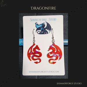 May include: Red dragon-shaped earrings with iridescent glitter accents. The earrings are displayed on a white card with the text "Dragonfire" and "Shimmerforge Studio". The earrings have silver-colored hooks.