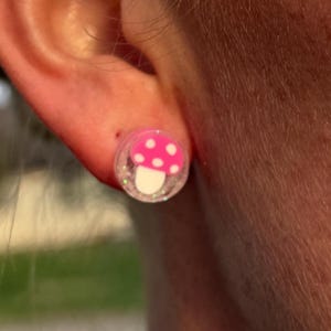 May include: A close-up of a person's ear with a pink mushroom-shaped earring. The earring has a white base with a pink mushroom cap and white polka dots. The earring is set in a clear, glittery material.