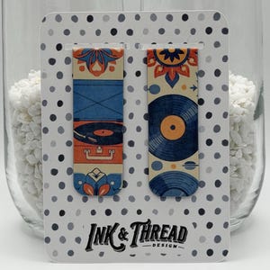 May include: Two decorative bookmarks with retro designs. One features a record player, the other a vinyl record. Both have blue, orange, and cream colours. The bookmarks are on a card with the text "Ink & Thread Design."