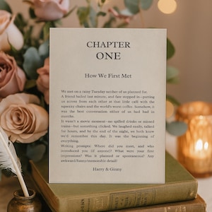 May include: A beige card with the text "CHAPTER ONE How We First Met" sits atop a stack of books. The card contains a handwritten story about a couple's first meeting. A quill and inkwell are in the foreground, with flowers in the background.