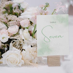 May include: A table number card with the word "Seven" in a soft green script, set against a watercolor wash. The card is displayed on a small wooden block, with a backdrop of white and pink flowers.