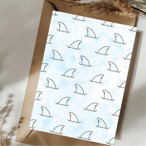 May include: A white card with a blue watercolor background and a repeating pattern of black shark fin outlines. The card is placed on a brown envelope.