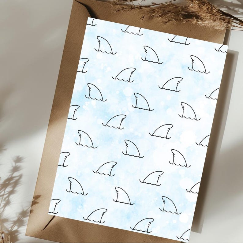 May include: A white card with a blue watercolor background and a repeating pattern of black shark fin outlines. The card is placed on a brown envelope.