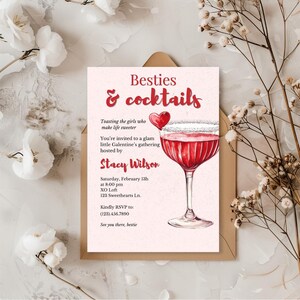 以下が含まれることがあります： 「Besties & cocktails」の赤い筆記体テキストが入ったピンク色の招待状。赤い飲み物で満たされたカクテルグラスの水彩イラストが特徴です。招待状は、Stacy Wilsonが主催するGalentineの集まり用です。