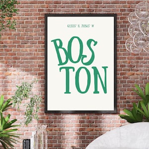 May include: A framed art print with the word "BOSTON" in large, green letters. Above the word is the text "42.3555 N, 71.0565 W". The print has a cream background and a black frame. The background is a red brick wall.