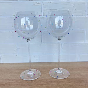 May include: Two clear wine glasses with long stems and round bases. The glasses are decorated with colorful polka dots. The glasses are on a wooden surface with a white brick wall in the background.